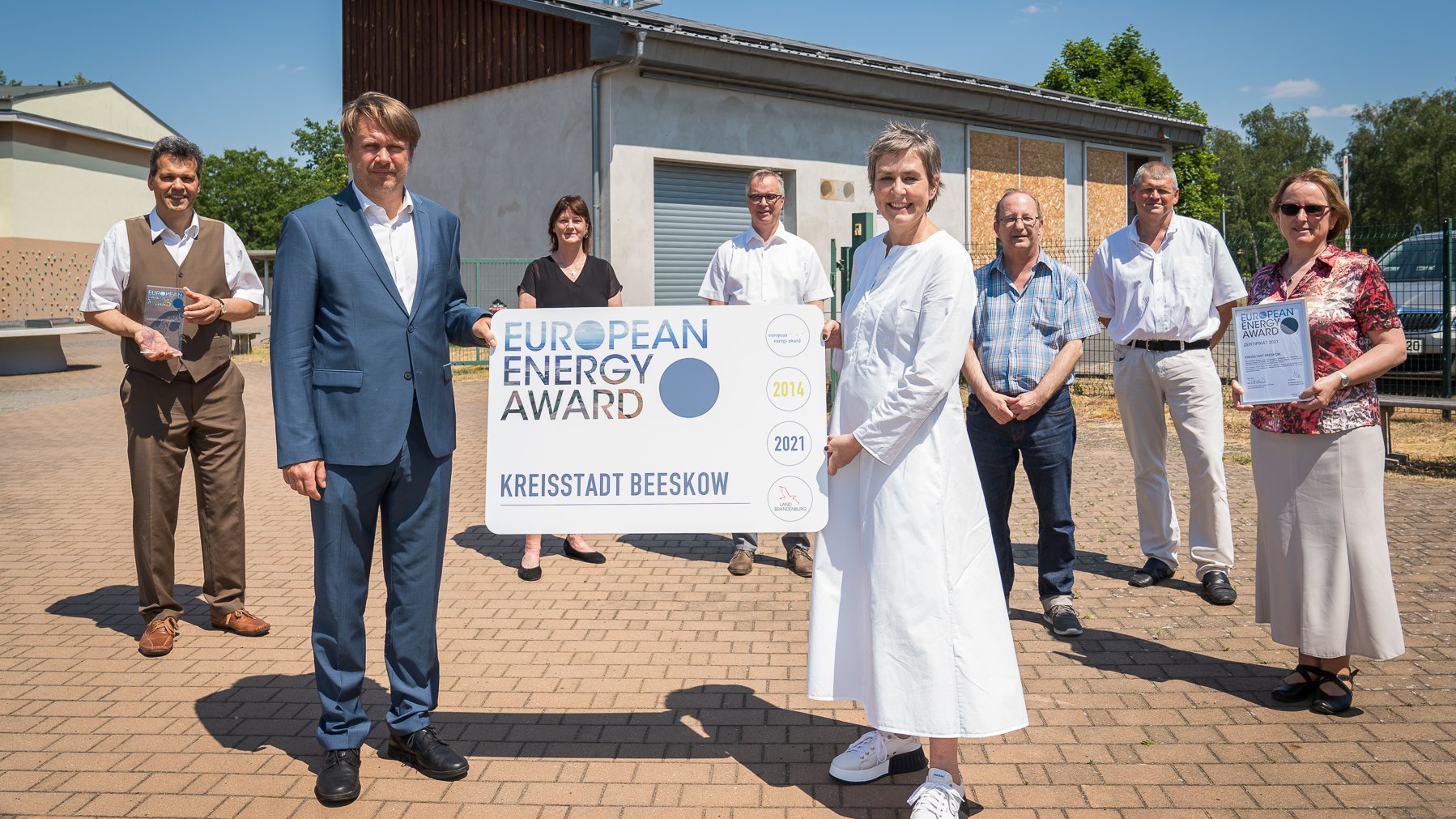 Drei Personen stehen im Freien auf gepflastertem Vorplatz; zwei Männer in Anzug und eine Frau in Business‑Kleidung halten ein großes Schild mit dem European Energy Award‑Logo und Text. Im Hintergrund ist eine moderne Fassade mit Glasfront und Besucherbereich erkennbar – Sinnbild für die Verleihung und das kommunale Energiemanagement der Stadt Beeskow