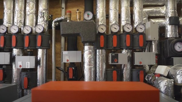 Nahaufnahme der Wärmepumpenanlage in der Verbandsgemeinde Liebenwerda: Zu sehen sind zwei graue, kompakte Wärmepumpen‑Innengeräte mit Anzeige‑Panels. Im Hintergrund sind dicke, glänzende Rohrleitungen zu erkennen, ebenso große Wärme‑ und Pufferspeicher (zylindrische Behälter). 