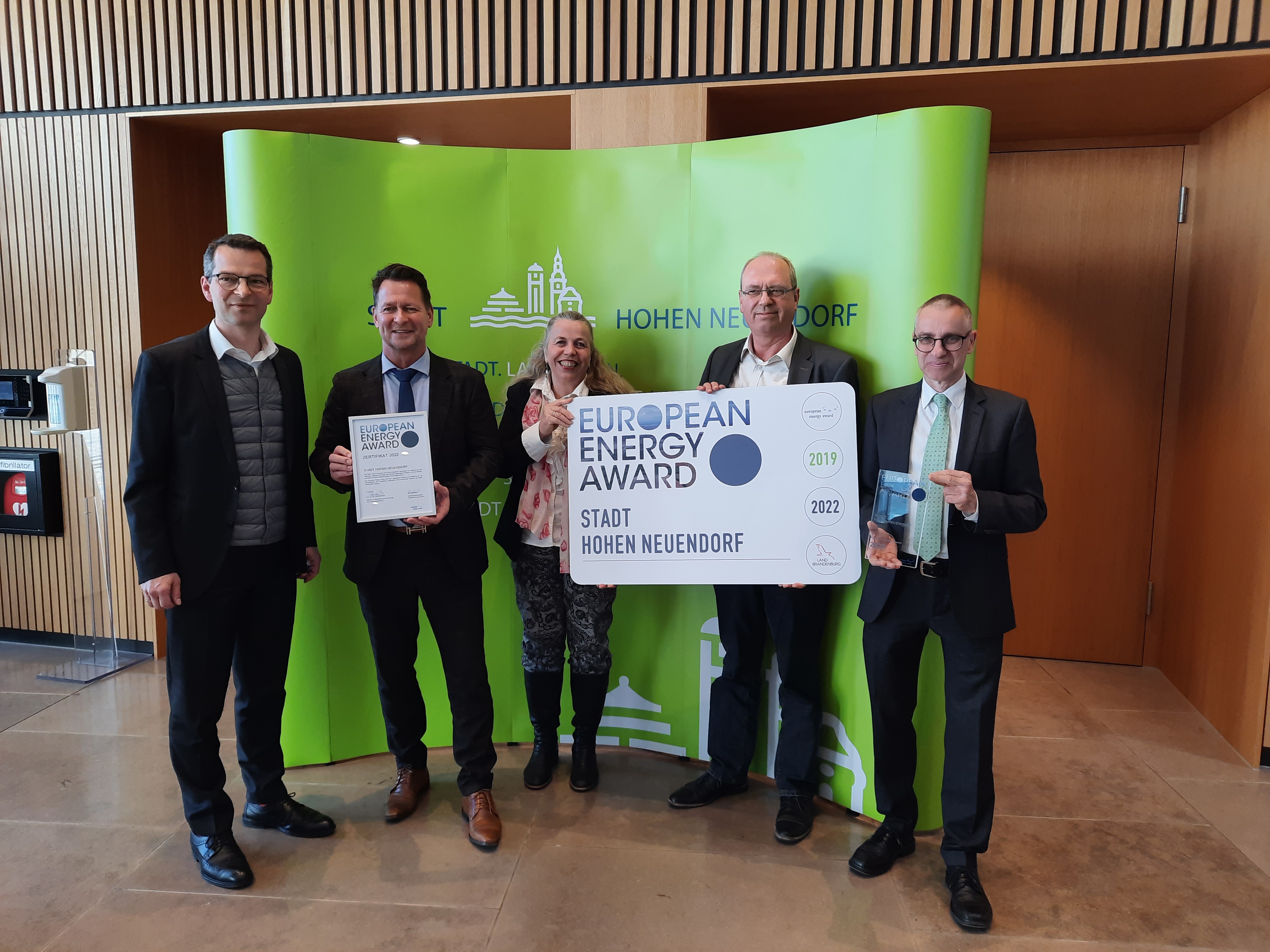 Verleihung des eea an Hohen Neuendorf. Die Preisträger halten eine Trophäe und ein Ortsschild des eea in der Hand. Auf dem Foto v.l.n.r. Hr. Saule, Geschäftsführer WFBB, Hr. Apelt Bürgermeister Hohen Neuendorf, Frau Ernst, Klimaschutzbeauftragte Hohen Neuendorf, Hr. Butz Bundesgeschäftsstelle eea, Dr. Weiland, Vorsitzender Stadtverordnetenversammlung